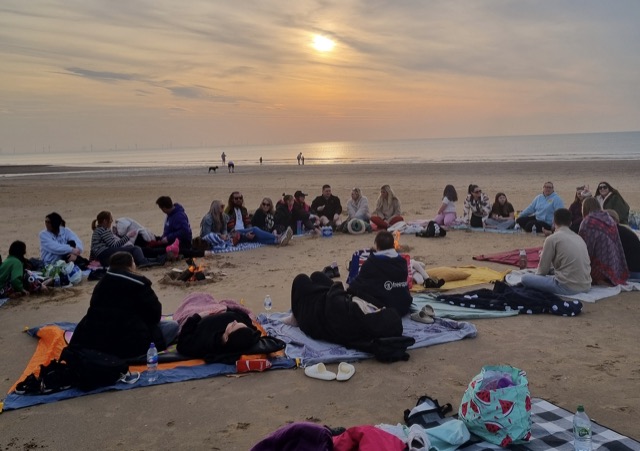 A circle of people sitting on the sand around a small fire as the sun sets over the sea.