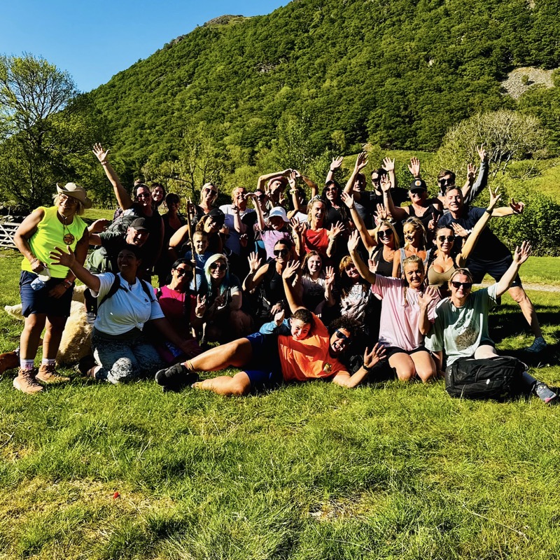 A large group of Nature Awareness walkers on a green hillside, arms raised and laughing.