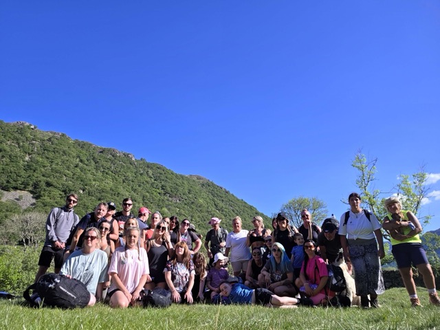 A large group posed on grass beneath a rocky hillside under a clear blue sky.