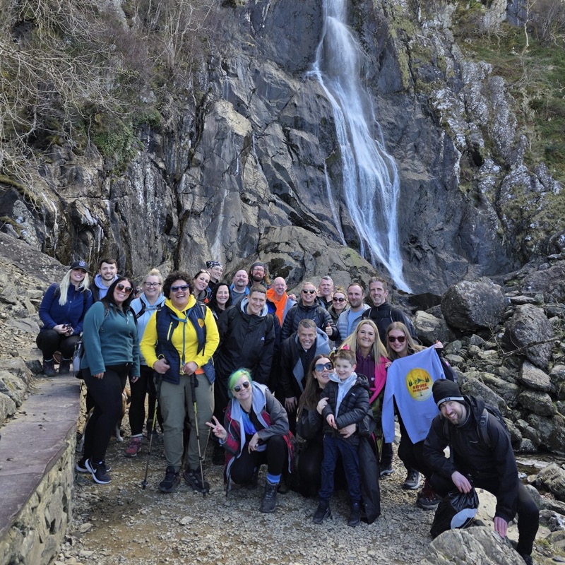 A Nature Awareness group gathered at the foot of a waterfall, one holding up a Nature Awareness t-shirt.