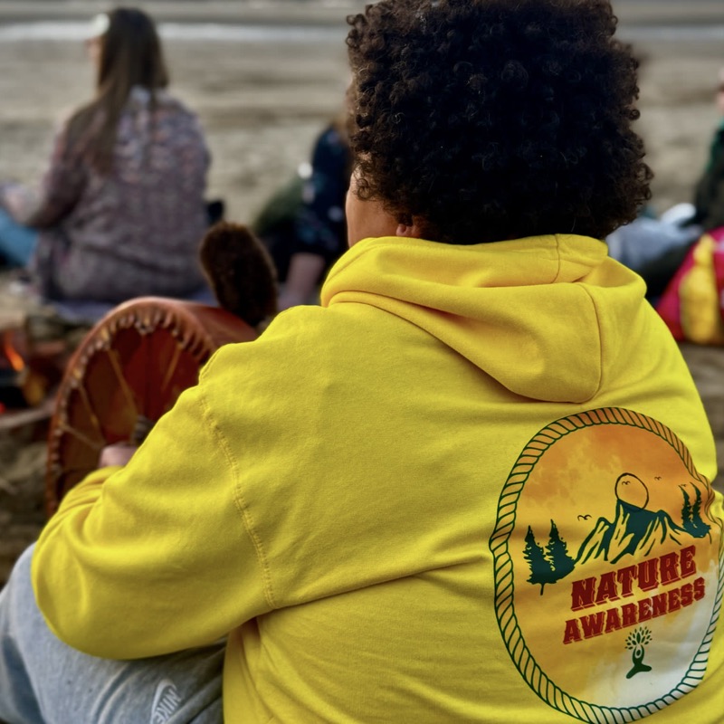 A participant in a Nature Awareness hoodie beside a shamanic drum at a beach fire, at dusk.