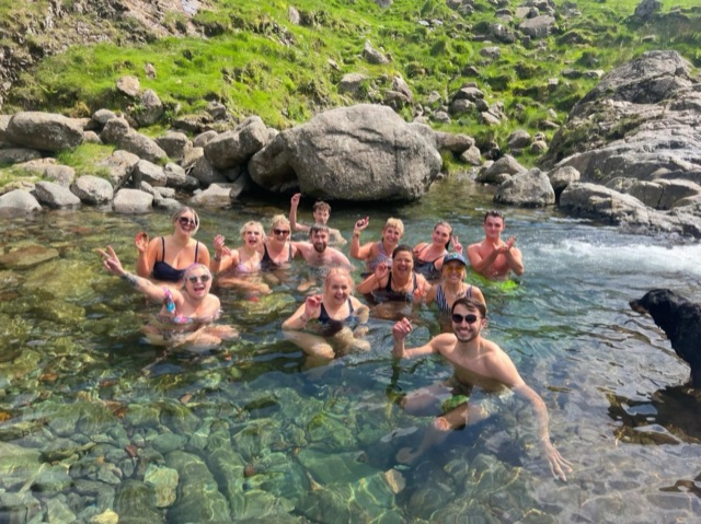 A group shoulder-deep in a clear river pool, laughing, with fells behind them.